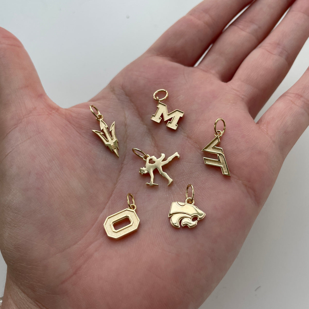 Michigan Logo Charm, Virginia Tech Logo Charm, Kansas State Wildcat Charm, Ohio State Logo Charm, ASU Pitchfork Charm, Wake Forest Walking Deacon Charm shown in gold in hand
