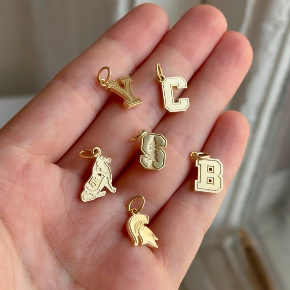 Five gold letter and mascot charms held in a person's hand against a soft neutral background.