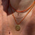 Close-up of a person wearing layered necklaces, including an orange cord necklace with a round gold pendant, a thin gold chain, and an orange beaded necklace, over a peach knit top.