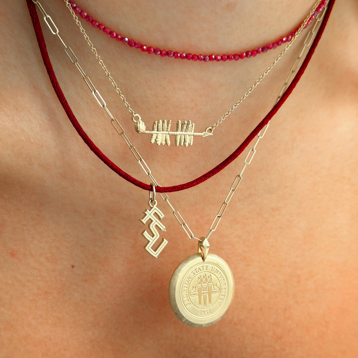 A close-up of a person's neck wearing a layered necklace set. The set includes a red beaded necklace, a red cord necklace with a gold arrow pendant, a silver paperclip chain with a gold 'ES' initial pendant, and a silver chain with a round gold University of State of Florida seal pendant.