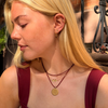 A close-up lifestyle shot of a blonde woman wearing layered gold necklaces, including a round coin pendant and a small bar necklace, along with a gold hoop earring, set against an outdoor brick wall background.
