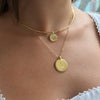 A close-up of a model wearing a layered gold necklace set, featuring a thin curved chain with a small round pendant and a longer chain with a larger round medallion pendant.