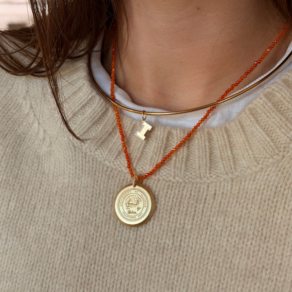 A close-up of a person wearing a layered necklace set. The set includes a gold bangle collar, a gold initial 'I' pendant, and a gold round medallion pendant on an orange cord, layered over a white shirt and a beige knit sweater.