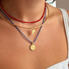Close-up of a person's neck and upper chest wearing three layered necklaces: a red beaded choker, a thin gold bangle, and a blue cord necklace with a small gold pendant and a larger round gold coin-style pendant.