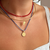 Close-up of a person's neck and upper chest wearing three layered necklaces: a red beaded choker, a thin gold bangle, and a blue cord necklace with a small gold pendant and a larger round gold coin-style pendant.