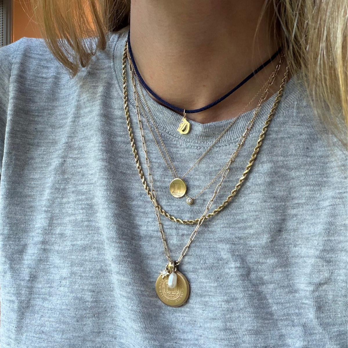 A close-up of a person wearing layered necklaces over a heather gray t-shirt. The necklaces include a dark navy blue cord choker with a small gold pendant, a gold chain with a round coin-style pendant, a twisted gold chain with a small disc and bead, and a longer gold chain with a textured round pendant featuring a small white pearl accent.