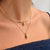 A close-up of a model's neck and upper chest wearing two layered gold necklaces: a thin chain necklace with a small Buddha head pendant and a slightly thicker curved chain necklace with a larger Buddha head pendant.