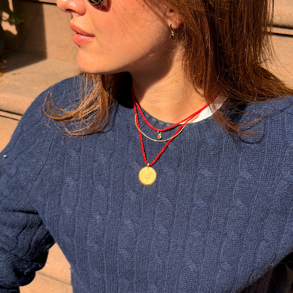 A woman wearing a navy blue cable-knit crewneck sweater, styled with light-wash blue jeans, a black belt, and layered red cord necklaces, sitting outdoors on stone steps.