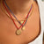 A close-up of a person's neck and chest wearing a layered necklace set. The set includes a gold bangle, a red cord necklace with a small gold bird charm, a blue cord necklace with a small round gold medallion, and a gold coin-style medallion on a red cord.
