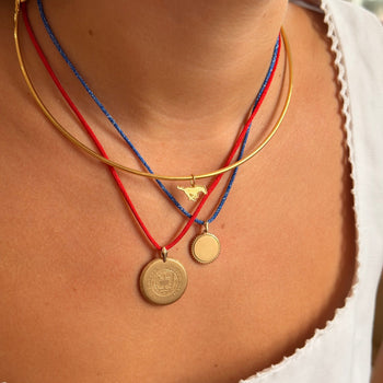A close-up of a person's neck and chest wearing a layered necklace set. The set includes a gold bangle, a red cord necklace with a small gold bird charm, a blue cord necklace with a small round gold medallion, and a gold coin-style medallion on a red cord.