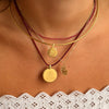 A close-up of a person's neck and upper chest wearing layered necklaces: a gold chain, a thin gold bangle, a red beaded necklace, and a red cord necklace with round gold pendants and a small gold leaf charm.