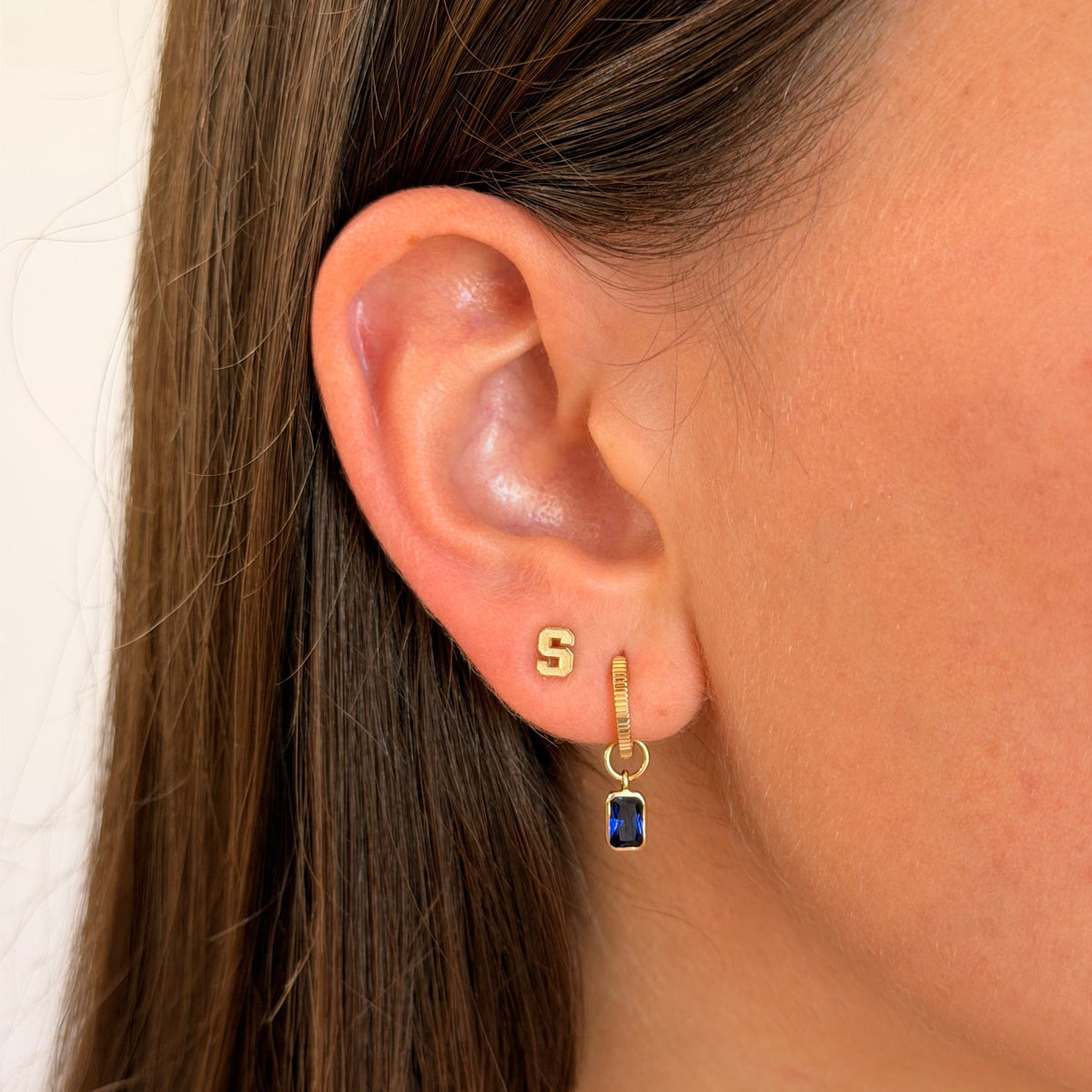 Close-up of a person's ear wearing a small gold hoop earring with a dangling gold huggie and a rectangular dark blue gemstone, against a light neutral background.