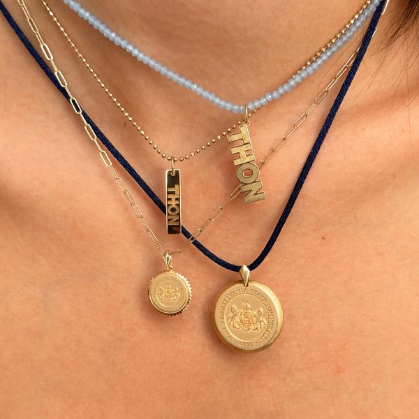 Layered necklaces worn on a person's neck, featuring gold-tone pendants including a baroque chain, a beaded light blue chain, a ball chain with a vertical 'THION' pendant, and two round gold medallion pendants on a dark cord.