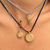 Layered necklaces worn on a person's neck, featuring gold-tone pendants including a baroque chain, a beaded light blue chain, a ball chain with a vertical 'THION' pendant, and two round gold medallion pendants on a dark cord.