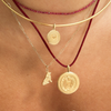 Layered necklaces worn on a model's neck, featuring a gold bangle, a beaded burgundy cord necklace, a silver chain necklace with a small dog charm, and a burgundy cord necklace with a large gold coin pendant.