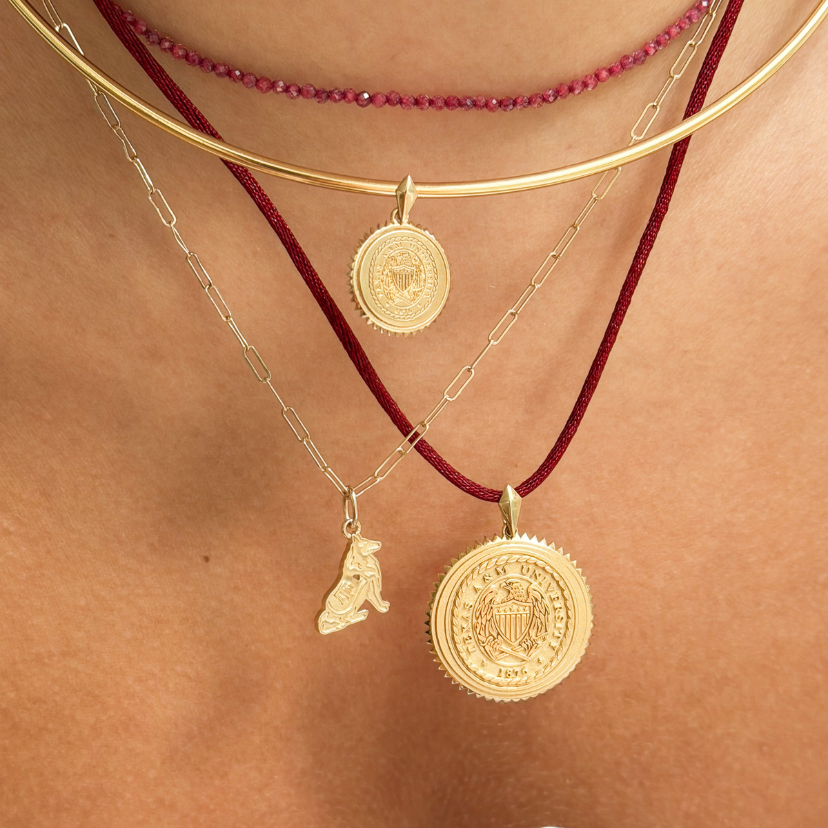 Layered necklaces worn on a model's neck, featuring a gold bangle, a beaded burgundy cord necklace, a silver chain necklace with a small dog charm, and a burgundy cord necklace with a large gold coin pendant.