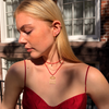 A blonde woman wearing a red spaghetti-strap top and layered gold necklaces, including a small bird-shaped pendant, standing outdoors in front of a brick building.