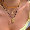 A close-up of a person's neck and upper chest wearing a layered necklace stack. The stack includes a thin gold chain, a textured orange beaded strand, a gold bangle, a black cord with a small gold geometric charm, a gold chain with a boot-shaped pendant, and a round gold coin pendant.